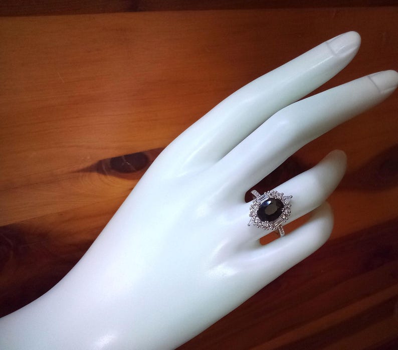 May include: A silver ring featuring an oval black gemstone, encircled by smaller, clear stones. The ring is displayed on a white hand, set against a wooden backdrop.