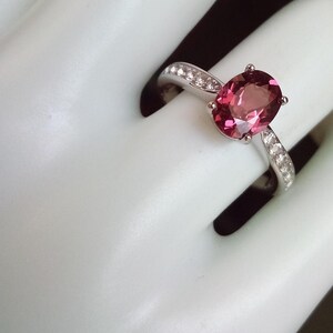 May include: A silver ring featuring a large, oval-cut, deep pink gemstone set in a four-prong mount. The band is crafted from polished silver and is accented with a row of small, clear, round-cut stones embedded along each side of the shank.