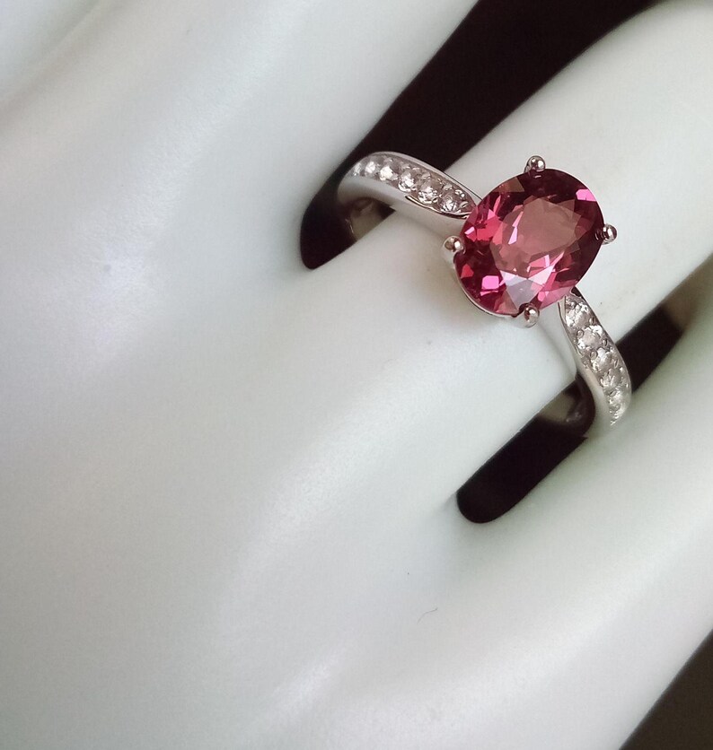 May include: A silver ring featuring a large, oval-cut, deep pink gemstone set in a four-prong mount. The band is crafted from polished silver and is accented with a row of small, clear, round-cut stones embedded along each side of the shank.