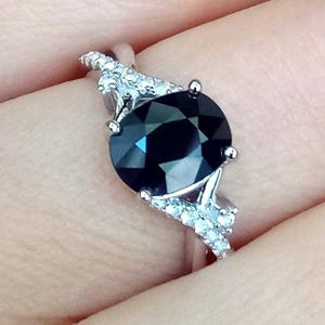 Puede incluir: Anillo de plata con una gran piedra negra ovalada y facetada. La banda est&aacute; adornada con piedras preciosas m&aacute;s peque&ntilde;as y transparentes. El dise&ntilde;o del anillo se destaca sobre un fondo neutro.
