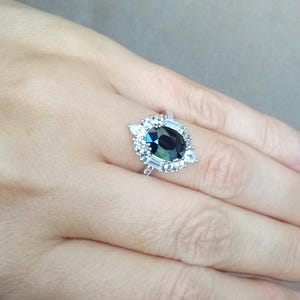 May include: A silver ring with a large, oval-cut, dark teal gemstone, encircled by smaller, clear, rectangular and triangular stones. The ring is displayed on a hand, highlighting the detailed design and sparkle of the jewellery.
