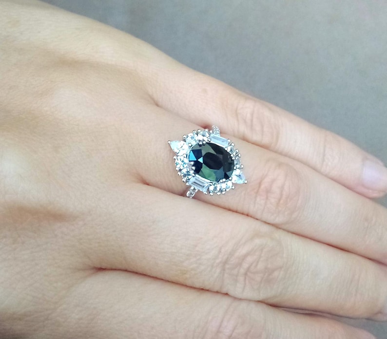 May include: A silver ring with a large, oval-cut, dark teal gemstone, encircled by smaller, clear, rectangular and triangular stones. The ring is displayed on a hand, highlighting the detailed design and sparkle of the jewellery.