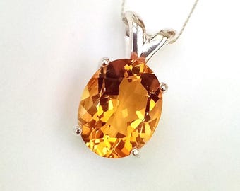 May include: A pendant necklace featuring an oval-cut, faceted golden-yellow gemstone set in a polished silver-toned metal prong setting. The gemstone displays a vibrant, translucent orange-yellow hue with internal light reflections.