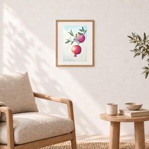 K&ouml;nnte beinhalten: Ein gerahmtes Aquarellbild, das zwei leuchtend rote Granat&auml;pfel zeigt, die an einem gr&uuml;nen, bebl&auml;tterten Zweig vor einem hellblauen Himmelshintergrund h&auml;ngen. Das Kunstwerk befindet sich in einem schlichten, hellen Holzrahmen.