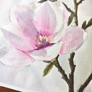 May include: A watercolor painting of magnolia flowers featuring soft pink and white petals with a yellow center. The blossoms are attached to dark brown, textured branches with small green leaves, rendered on a white paper background.