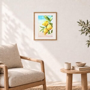 May include: A framed watercolour print featuring three bright yellow lemons hanging from a green leafy branch. The artwork is set against a soft, blended background of light blue at the top and a warm, pale peach tone at the bottom, encased in a light wood frame.