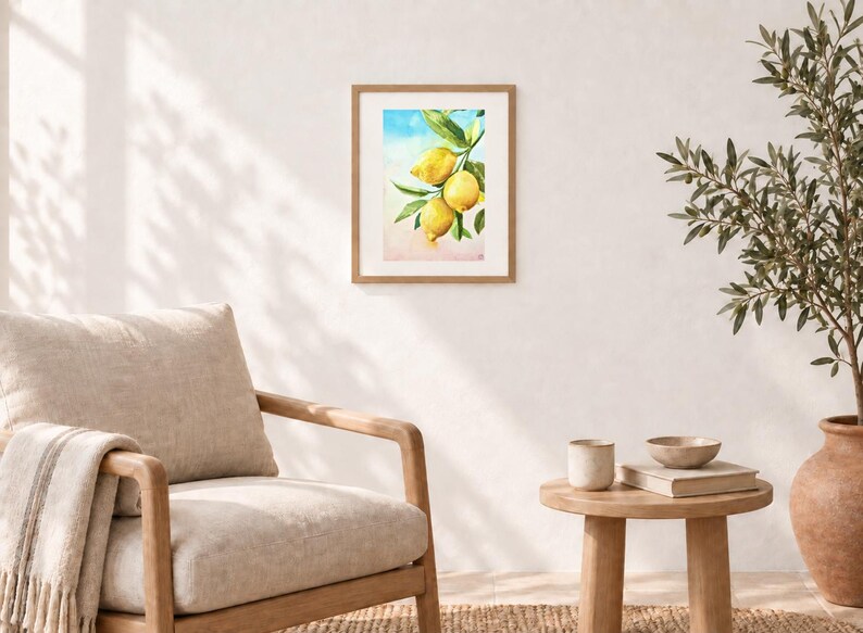 May include: A framed watercolour print featuring three bright yellow lemons hanging from a green leafy branch. The artwork is set against a soft, blended background of light blue at the top and a warm, pale peach tone at the bottom, encased in a light wood frame.