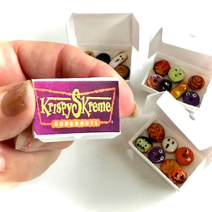 May include: Miniature white cardboard boxes containing small, colorful, themed doughnut-shaped charms. One box features a purple and orange label with the text Krispy Skreme Doughnuts. The charms include various faces, patterns, and colors.