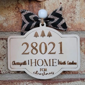 Op de afbeelding: Houten kerstversiering met het adres 28021, het woord "HOME" en de tekst "Cherryville, North Carolina". De versiering heeft een zwart-beige strik en een witte kraal.