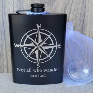 Könnte beinhalten: Schwarze Flasche mit Kompassrosen-Design und dem Zitat "Not all who wander are lost". Die Flasche hat einen schwarzen Deckel und ein kleines Glas. Neben der Flasche befindet sich ein weißer Netzbeutel.