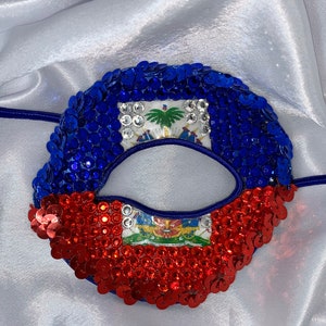 Puede incluir: Una máscara de ojos de lentejuelas azules y rojas con el diseño de la bandera haitiana. La máscara tiene una pequeña abertura para los ojos.