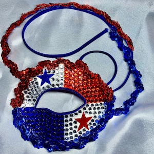 May include: A patriotic mask with red, white, and blue sequins and star accents. The mask is attached to a blue elastic band and a sequined headband. The design is reminiscent of a flag.