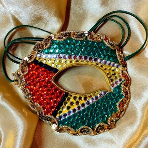 May include: A masquerade mask featuring a vibrant design of red, yellow, green, black, and white rhinestones arranged in a geometric pattern. The mask is bordered by gold sequins and includes thin green elastic bands for securing it in place.