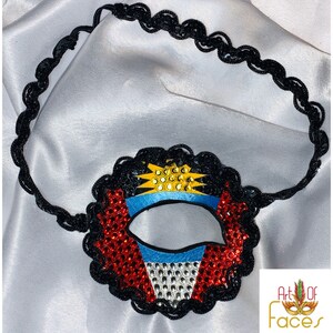 May include: A black eye mask with a red, white, and blue design featuring a yellow sun with a blue and white background. The mask is decorated with rhinestones and has a black elastic band.