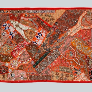 May include: A colorful patchwork wall tapestry with a red border. The tapestry is made of various fabrics and is decorated with beads, sequins, and embroidery. The tapestry features a variety of patterns and colors, including orange, brown, pink, and green.