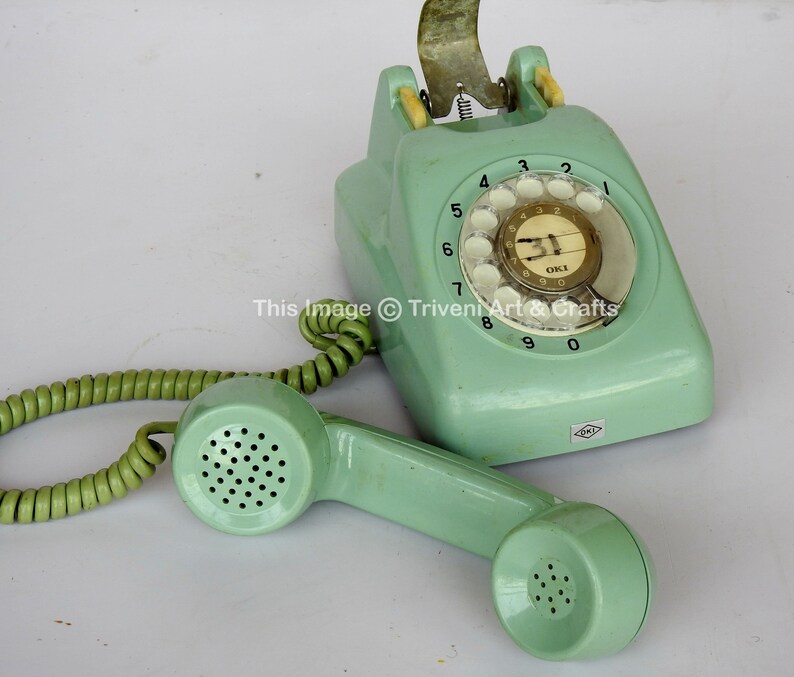 May include: A vintage green rotary dial phone with a coiled cord and a handset. The phone has a white dial with black numbers and a white label with the text "OKI" on it.