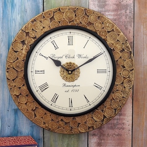 Könnte beinhalten: Eine runde Wanduhr in Goldtönen mit römischen Ziffern und einem schwarzen Zifferblatt. Das Zifferblatt der Uhr zeigt "Royal Clock Works Birmingham est. 1792". Die Uhr ist von einem dekorativen Rand aus goldfarbenen Münzen umgeben.