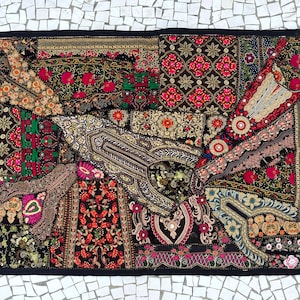 Op de afbeelding: Een rechthoekig patchwork textiel met een zwarte rand. Het ontwerp bevat een verscheidenheid aan patronen in tinten rood, roze, groen, goud en zwart. Bloemen- en geometrische motieven zijn prominent aanwezig.