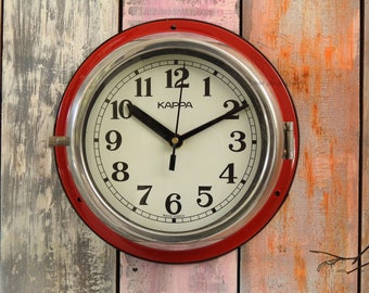 May include: A red and silver round wall clock with black hands and black numbers. The clock face is white with the word "KAPPA" printed in black. The clock is made in Korea.