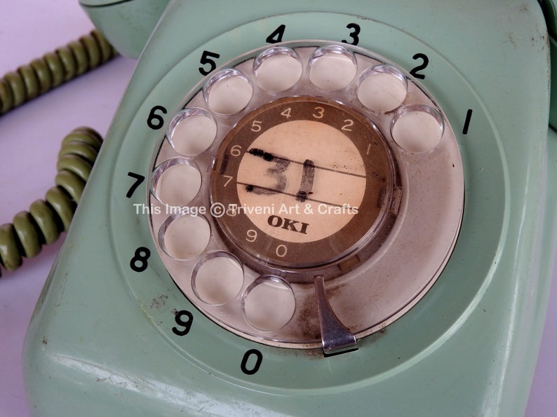 May include: A vintage green rotary dial phone with a white dial and black numbers. The phone has a silver push button and the text "OKI" is visible on the dial.