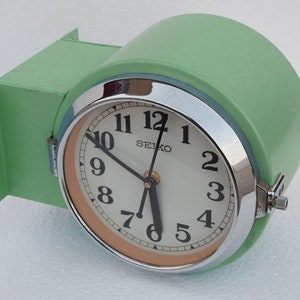 Op de afbeelding: Een groene wandklok met een witte wijzerplaat en zwarte wijzers. De klok heeft een chromen rand en een binnenring in perzikkleur. De wijzerplaat van de klok heeft "SEIKO" en de cijfers 1 tot en met 12.