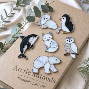 May include: Six stained glass figures of Arctic animals including a seal, penguin, orca, fox, owl, and polar bear. The figures are arranged on a tan surface with the text Arctic animals Printable patterns printed in black at the bottom.