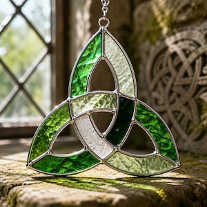 May include: A stained glass Celtic knot suncatcher. The design features a three-part knot in shades of green and white, framed by a silver metal border. The suncatcher hangs from a silver chain, and is displayed in front of a window.