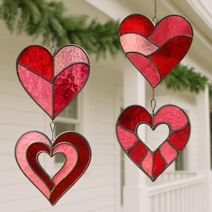 Pode incluir: Quatro ornamentos de vidro manchado em forma de coração em tons de vermelho e rosa, pendurados em correntes. Cada coração tem um design diferente, com padrões e texturas variadas. Os ornamentos são exibidos ao ar livre.