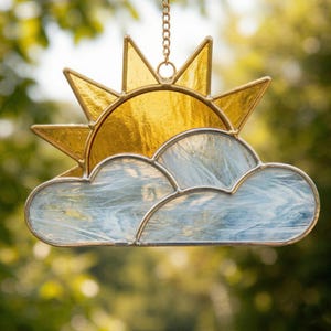 Può includere: Decorazione a sospensione con sole e nuvola in vetro colorato. Il sole è giallo brillante con raggi appuntiti e la nuvola è azzurra con vortici bianchi. Il sole e la nuvola sono collegati con una catena dorata.