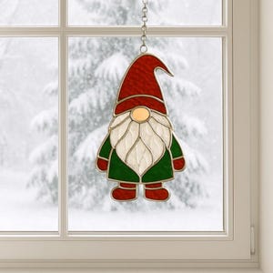 Op de afbeelding: Een gebrandschilderd glazen raamdecoratie van een kabouter. De kabouter heeft een rode hoed, een witte baard en een groene jas. Hij hangt voor een raam met een besneeuwde achtergrond aan een zilveren ketting.