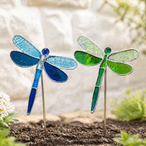 Pode incluir: Duas decorações de jardim em forma de libélula. Uma libélula é azul, com tons de azul claro e escuro. A outra é verde, com tons de verde claro e escuro. Ambas têm molduras de metal prateado e estão plantadas no solo.