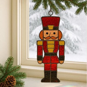 Puede incluir: Un adorno navideño de vidriera con forma de soldado cascanueces. El adorno presenta un uniforme rojo con detalles dorados, botas negras y un sombrero alto rojo. Cuelga de un anillo de metal, con una escena invernal nevada de fondo.
