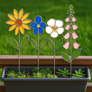 May include: A collection of colorful stained glass flower garden stakes. The stakes feature a yellow sunflower, a blue flower, a white flower, and a pink foxglove. The flowers are set in a black planter box with green plants.