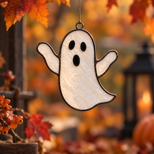 Puede incluir: Un adorno de fantasma de vitral con un cuerpo blanco, texturizado y translúcido, con contornos de vidrio negro para los ojos y la boca. El fantasma tiene dos brazos levantados y un borde inferior curvo, suspendido por un hilo fino.