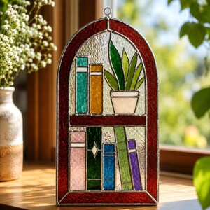 Puede incluir: Vidriera que representa una estanter&iacute;a con libros coloridos y una planta en maceta. El marco es de color rojo oscuro y el vidrio tiene textura. Los libros son de varios colores, incluyendo verde, amarillo, azul y morado. Una pieza decorativa para la decoraci&oacute;n del hogar.