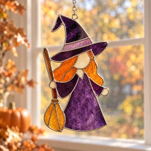 May include: A stained glass suncatcher shaped like a witch wearing a purple hat and dress, featuring orange hair and holding a yellow broomstick. The piece is constructed with dark metal lead lines and translucent glass in shades of purple and orange.