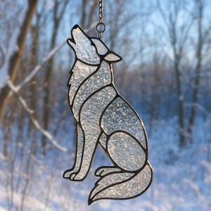 May include: A stained glass howling wolf suncatcher. The wolf is depicted in a sitting position with its head raised and mouth open. The glass is textured and clear, with black outlines. The suncatcher hangs from a chain against a blurred winter background.
