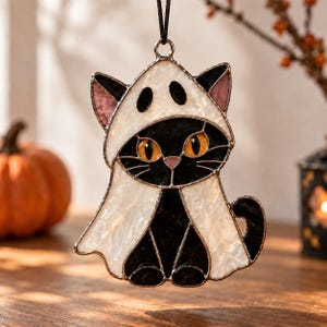 Könnte beinhalten: Ein Buntglas-Suncatcher in Form einer schwarzen Katze, die ein weißes Geisterkostüm trägt. Die Katze hat leuchtend orangefarbene Augen, eine rosa Nase und schwarze Schnurrhaare. Das weiße Glas hat eine texturierte Perlmuttoptik und hängt an einer schwarzen Schnur.