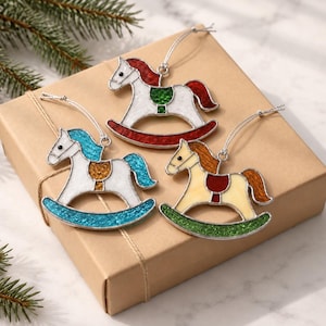 Peut inclure: Trois décorations en verre teinté en forme de cheval à bascule avec des contours en métal argenté et des cordons de suspension. Un cheval est blanc avec une crinière et un socle rouges, un bleu, et un crème avec une crinière orange et un socle vert.
