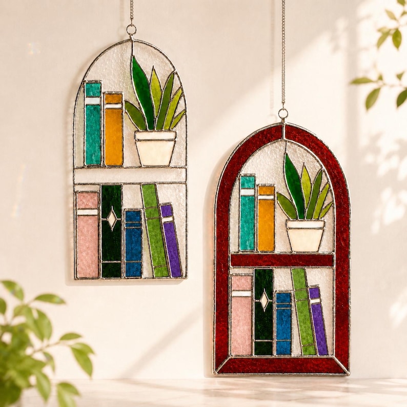 Puede incluir: Dos decoraciones de estanter&iacute;a de vidrieras. Una tiene un marco plateado, la otra un marco rojo. Cada una presenta coloridos libros de vidrio y una planta en maceta. Los libros son en tonos de rosa, verde, azul, naranja y morado. Las plantas tienen hojas verdes.