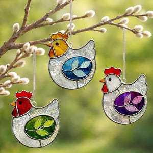 May include: Three stained glass chicken ornaments hanging from a tree branch. Each chicken has a different colored comb and wattle, with a colorful egg-shaped design on the body. The chickens are clear with a textured appearance.