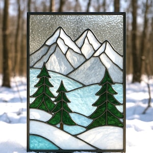 May include: Stained glass panel featuring a winter mountain scene. The artwork depicts snow-covered mountains, evergreen trees, and a snowy landscape in shades of white, blue, and green, framed by a black border. The panel is rectangular.