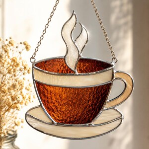 May include: A stained glass suncatcher shaped like a coffee cup on a saucer with rising steam. The cup features textured amber glass, a cream-colored rim and handle, and translucent white steam, all held together by metal solder and a hanging chain.