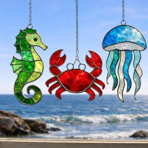 May include: Three stained glass sea creatures hang from chains: a green seahorse, a red crab, and a blue and white jellyfish. The ocean and sky are visible in the background. These decorative pieces are perfect for a coastal theme.