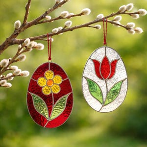 Könnte beinhalten: Zwei Ostereier-Ornamente aus Glasmalerei, die an einem Ast hängen. Ein Ei zeigt ein gelbes Blumendesign auf rotem Hintergrund, das andere eine rote Tulpe auf weißem Hintergrund. Beide sind silberfarben umrandet und haben rote Aufhängeschnüre.