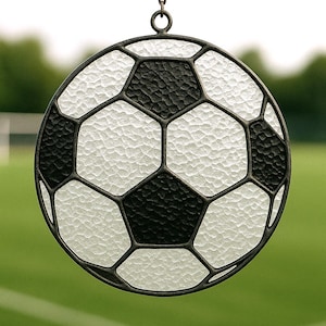 Peut inclure: Ornement rond en vitrail représentant un ballon de football. Le motif présente des pentagones noirs et blancs, encadrés par un bord en métal foncé. Il est suspendu à une chaîne sur un fond vert flou, évoquant le thème du sport.