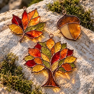 May include: Three stained glass suncatchers featuring autumn themes: a maple leaf, an acorn, and a tree. Each piece is crafted with multicoloured glass in shades of red, orange, yellow, and green, held together by dark metal solder lines with loops.