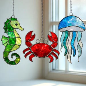 May include: Three stained glass sea creatures hang from chains: a green seahorse, a red crab, and a blue jellyfish. The seahorse has a yellow head and green body. The crab is red. The jellyfish is blue and white with flowing tentacles.
