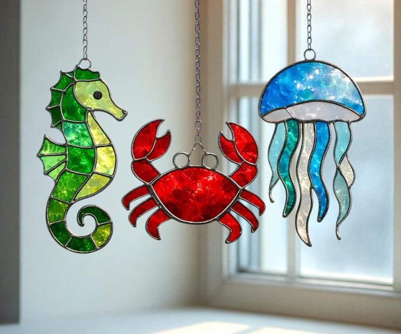 May include: Three stained glass sea creatures hang from chains: a green seahorse, a red crab, and a blue jellyfish. The seahorse has a yellow head and green body. The crab is red. The jellyfish is blue and white with flowing tentacles.