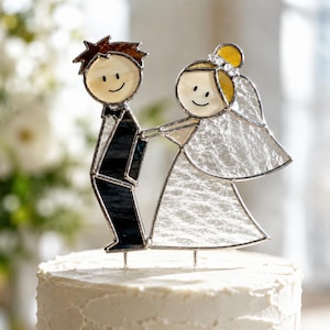 May include: A stained glass cake topper featuring two stylized figures. One figure wears a dark suit with a black bowtie, while the other wears a white dress and a textured veil. Both figures have cream-colored faces with simple black line features.